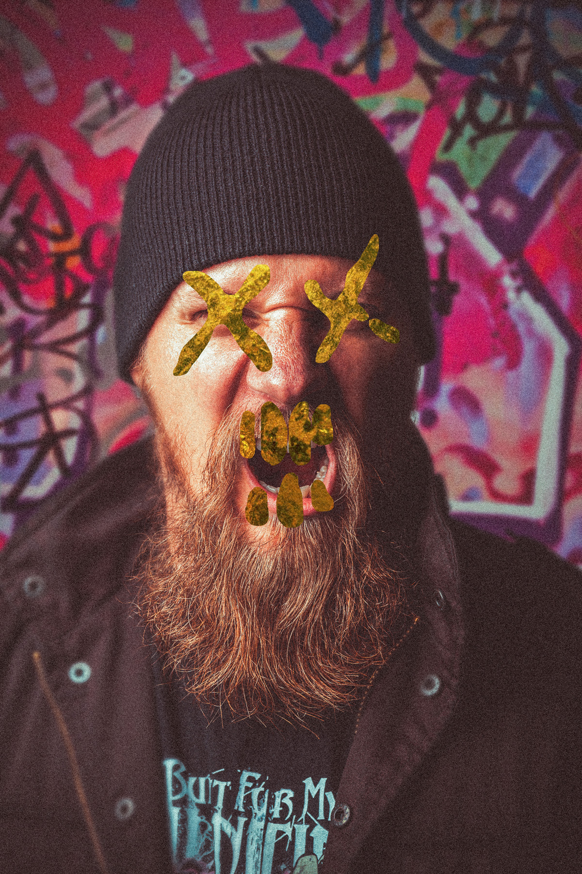 A gritty urban portrait of a bearded man in a black beanie and jacket, shouting with a stylized gold skull and cross mask painted over his face, set against a colorful graffiti-covered wall.